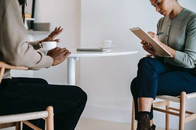 A man talks to a woman taking notes, both sitting at a table.